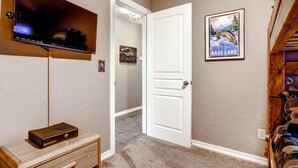 A hallway with a flat-screen TV mounted on the wall, a wooden cabinet with a stereo system, a door leading to another room, and a framed picture of Bass Lake. - Anglers Rest (Oakhurst)
