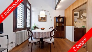 Un comedor con una mesa redonda, dos sillas, una lámpara y una estantería. Se observan dos ventanas con vidrio decorativo, un radiador y una cocina al fondo.