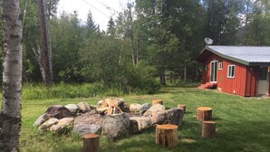 A rustic cabin with a red exterior and a satellite dish on the roof, surrounded by a grassy area with a pile of firewood and rocks. - Peacful and cozy cottage in the mountains (Bigfork)