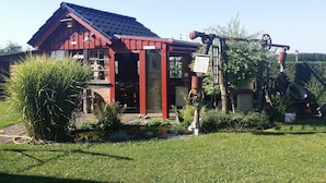 A red wooden house with a sloped roof, a small garden with a pond, and a wheelbarrow.