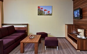A modern hotel room with a sofa, a wooden table, a small ottoman, a painting of red flowers, a television, and a minibar. - Alexandra Business Hotel (Puchov)