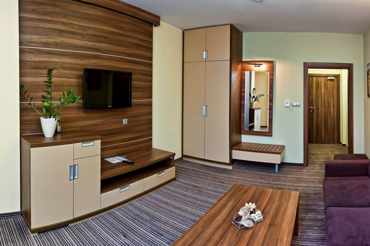 A modern hotel room with a wooden accent wall, a flat-screen TV, a sofa, a coffee table, and a bed with a headboard.