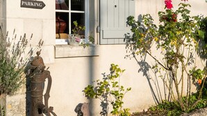 A building with a 'PARKING' sign, a window with blue shutters, and a small garden with plants and a decorative metal structure. - Le Clos de La Rousselière B&B (Francueil)