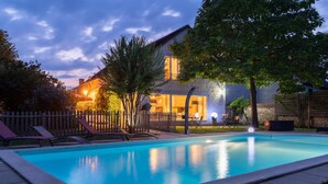 A swimming pool with lounge chairs, a wooden fence, and a house with a lit interior. - Le Clos de La Rousselière B&B (Francueil)