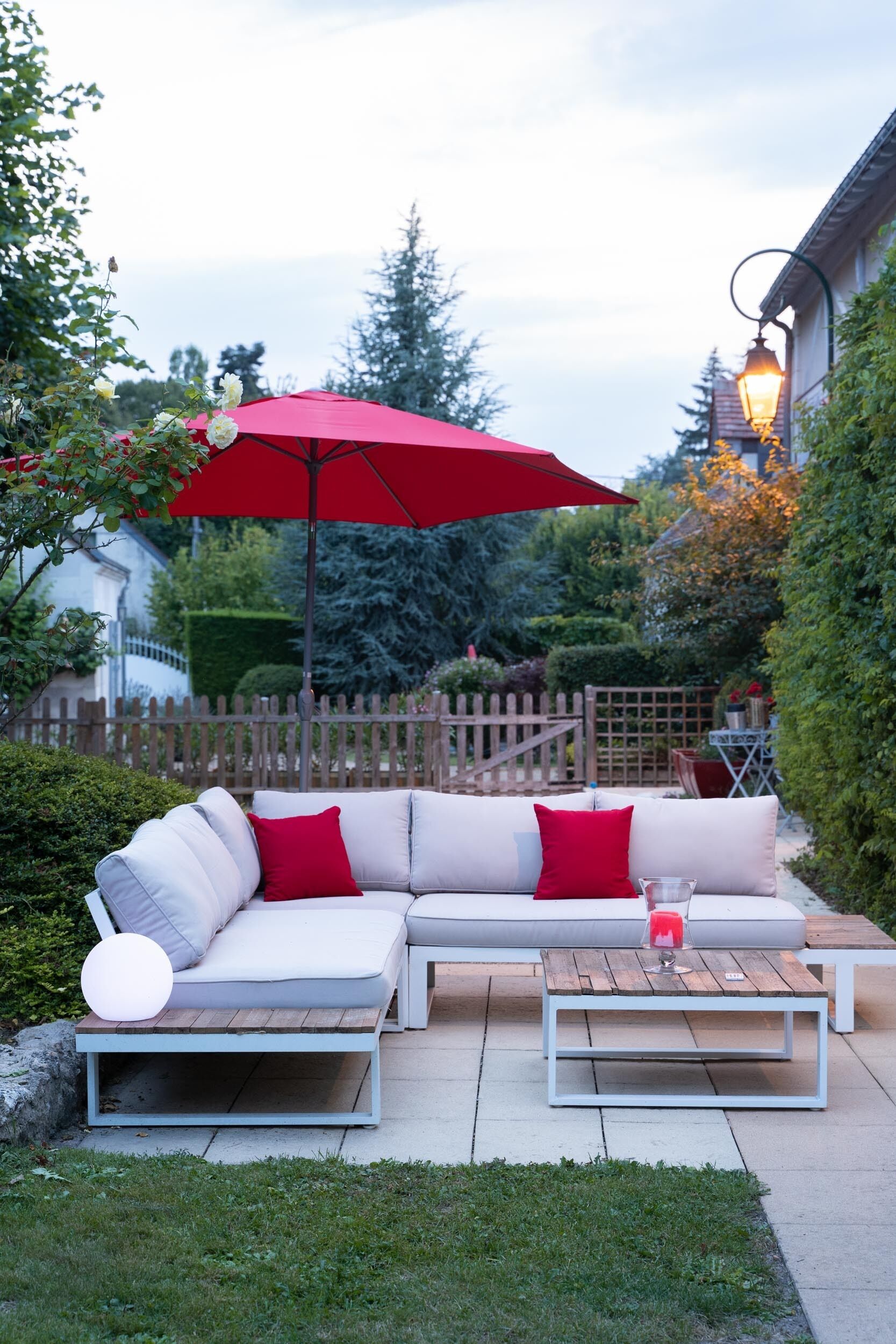 An outdoor patio with a white sectional sofa, red cushions, a wooden coffee table, and a red umbrella.