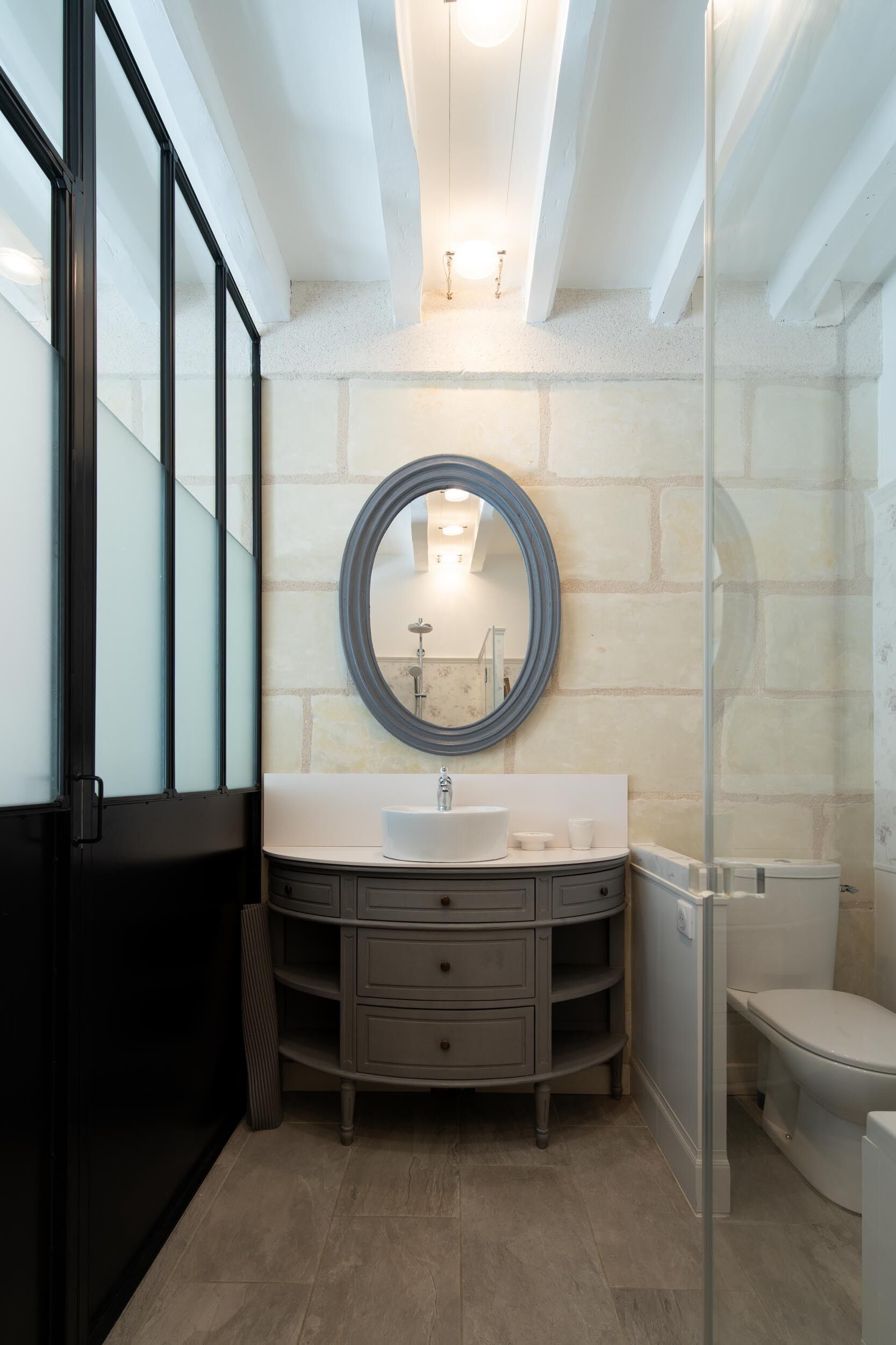 A modern bathroom with a glass shower enclosure, a round mirror, and a vanity with a sink and drawers.