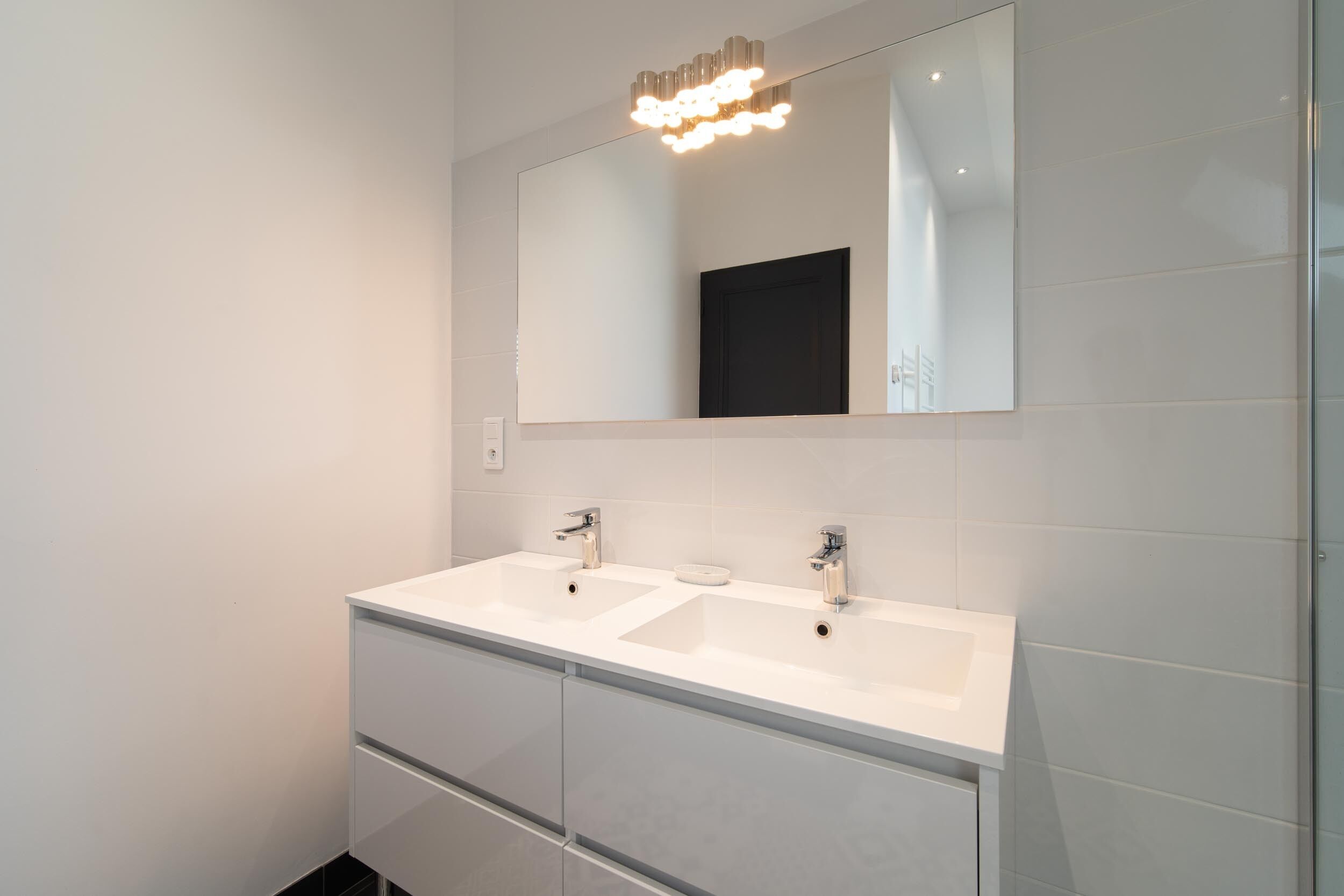 A modern bathroom with two sinks, a large mirror, and a contemporary light fixture.