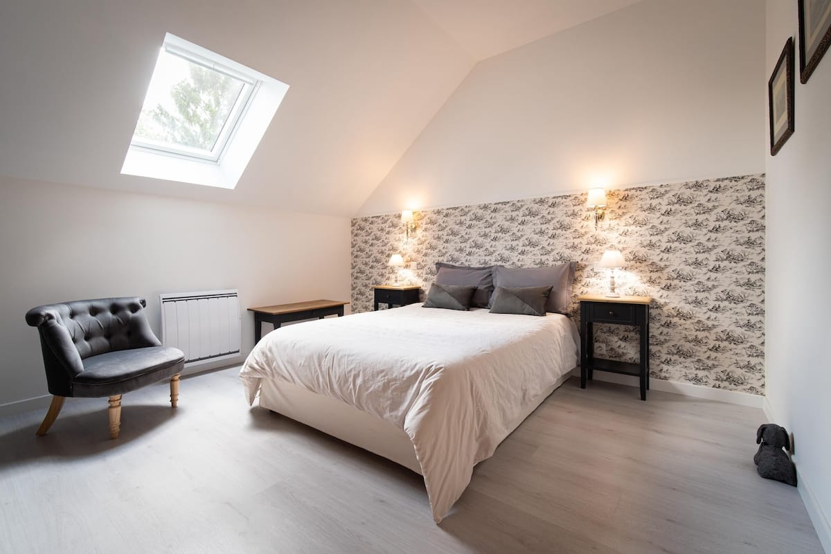 Ein Schlafzimmer mit einem Bett, einem Sessel, zwei Nachttischen, einer Wandleuchte und einem Fenster.