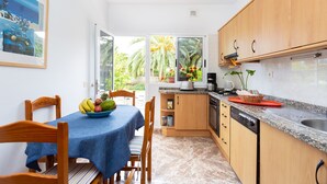 A compact kitchen with wooden cabinets, a dining table with a blue tablecloth, and a view of a tropical garden through the window. - Finca La Gaviota - Los Limoneros (Icod de los Vinos)