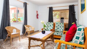 A room with a wooden table, wicker chairs, a sofa with plaid cushions, and a view of a garden through a window. - Finca La Gaviota - Los Limoneros (Icod de los Vinos)