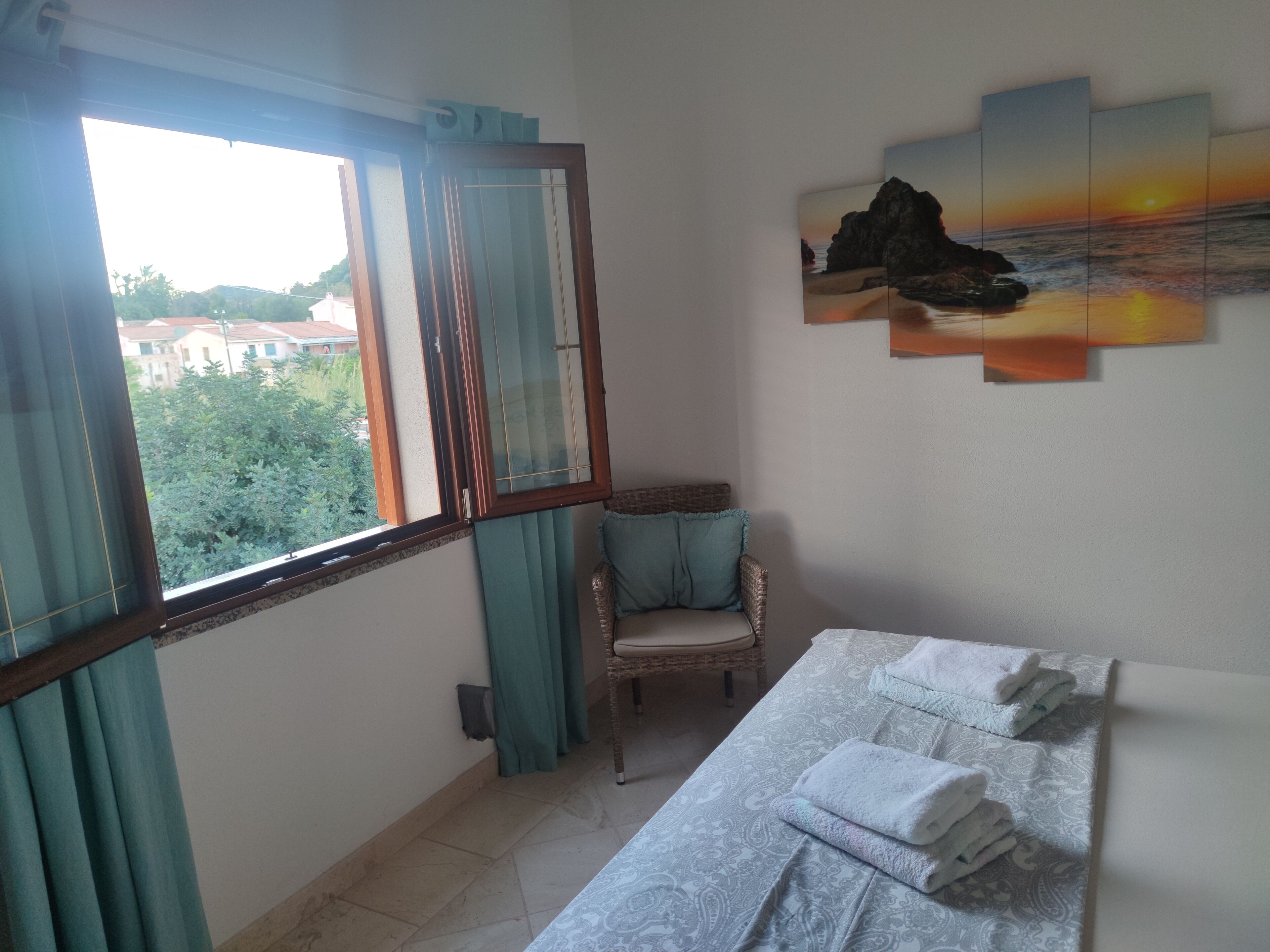Una habitació amb llit, cadira, finestra amb cortines i una obra d'art penjada a la paret.
