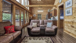 A spacious living room with a large brown leather sofa, a smaller sofa with red and blue cushions, wooden walls, large windows, and a chandelier. - Secluded cabin-Movie theater, outdoor kitchen and outdoor tv- Mountain View-gameroom (SEVIERVILLE)