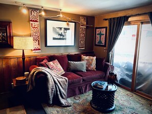A cozy living room with a red sofa, patterned cushions, a coffee table, and a window with a view.