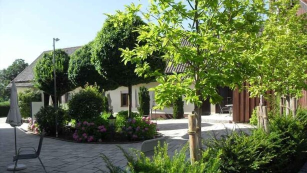 Un cortile con pavimento in piastrelle, circondato da vegetazione lussureggiante e un edificio con tetto in tegole.
