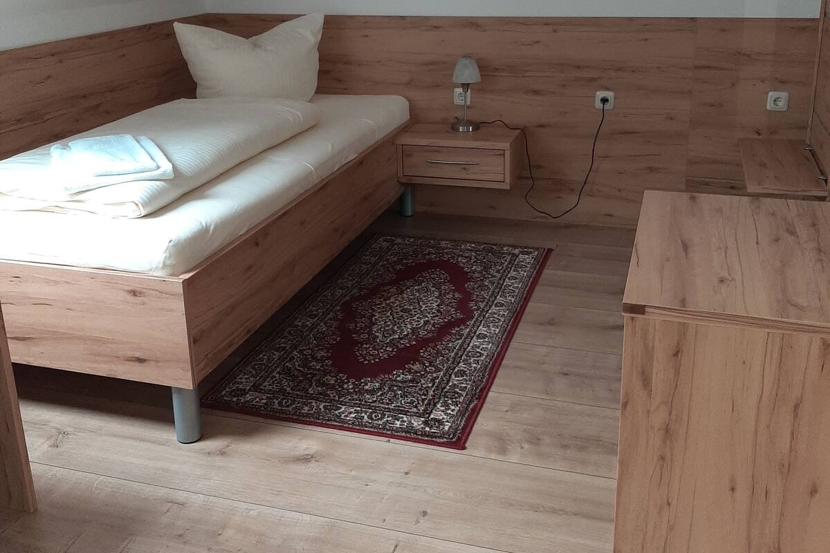 A bedroom with a wooden bed, a nightstand with a lamp, a red patterned rug, and a small wooden dresser.