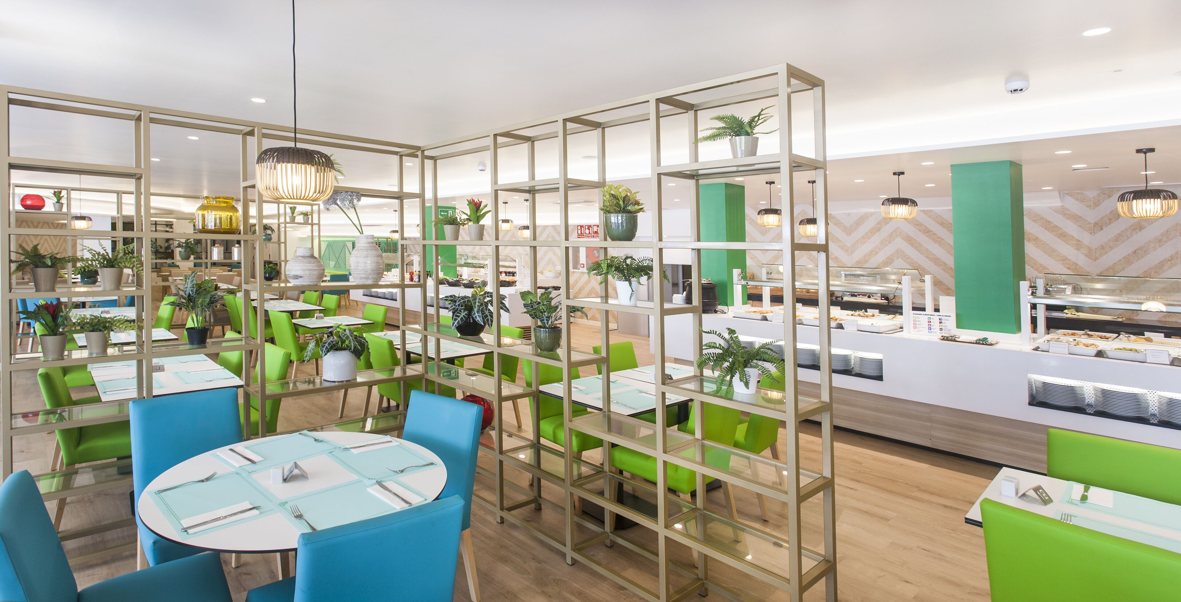 Un restaurante moderno con asientos verdes y azules, separaciones de vidrio y plantas en macetas.