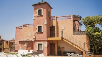 Un bâtiment à la façade rose, doté d’un balcon et d’un escalier menant à un étage supérieur. On aperçoit des fauteuils de jardin au premier plan.