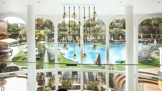 A pool area with lounge chairs and umbrellas, surrounded by greenery and a view through large windows. - Hotel Villa Mandi Golf Resort (Arona)