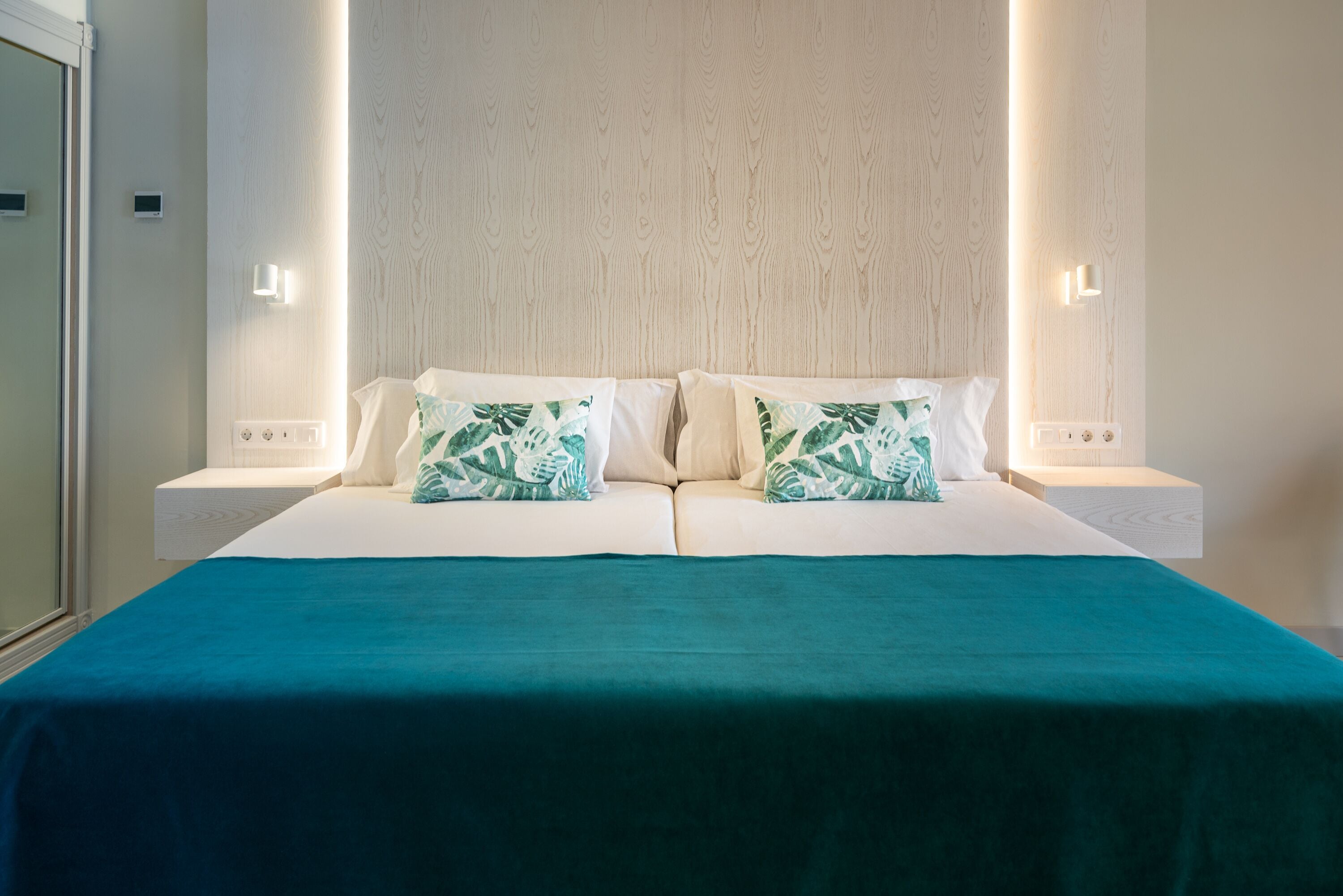 A neatly made double bed with a teal bedspread and two pillows with a leaf pattern, flanked by nightstands with wall-mounted lights.