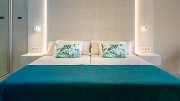 A neatly made double bed with a teal bedspread and two pillows with a leaf pattern, flanked by nightstands with wall-mounted lights.