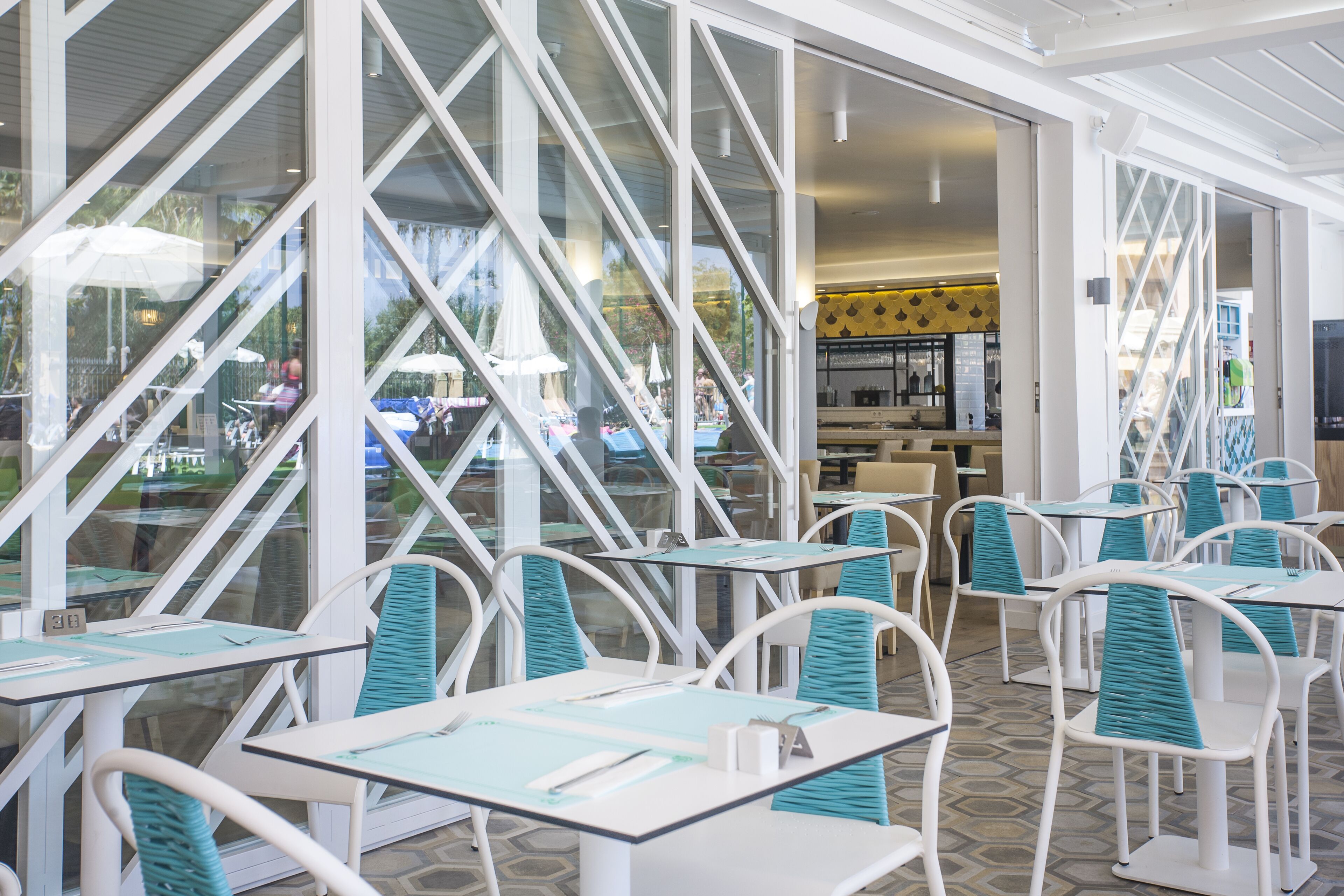 A restaurant with white tables and chairs, large windows, and a view of the outside.