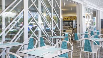 A restaurant with white tables and chairs, large windows, and a view of the outside.