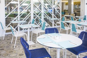 Un restaurante con mesas blancas y sillas azules, un suelo estampado y amplios ventanales que ofrecen vistas al exterior.