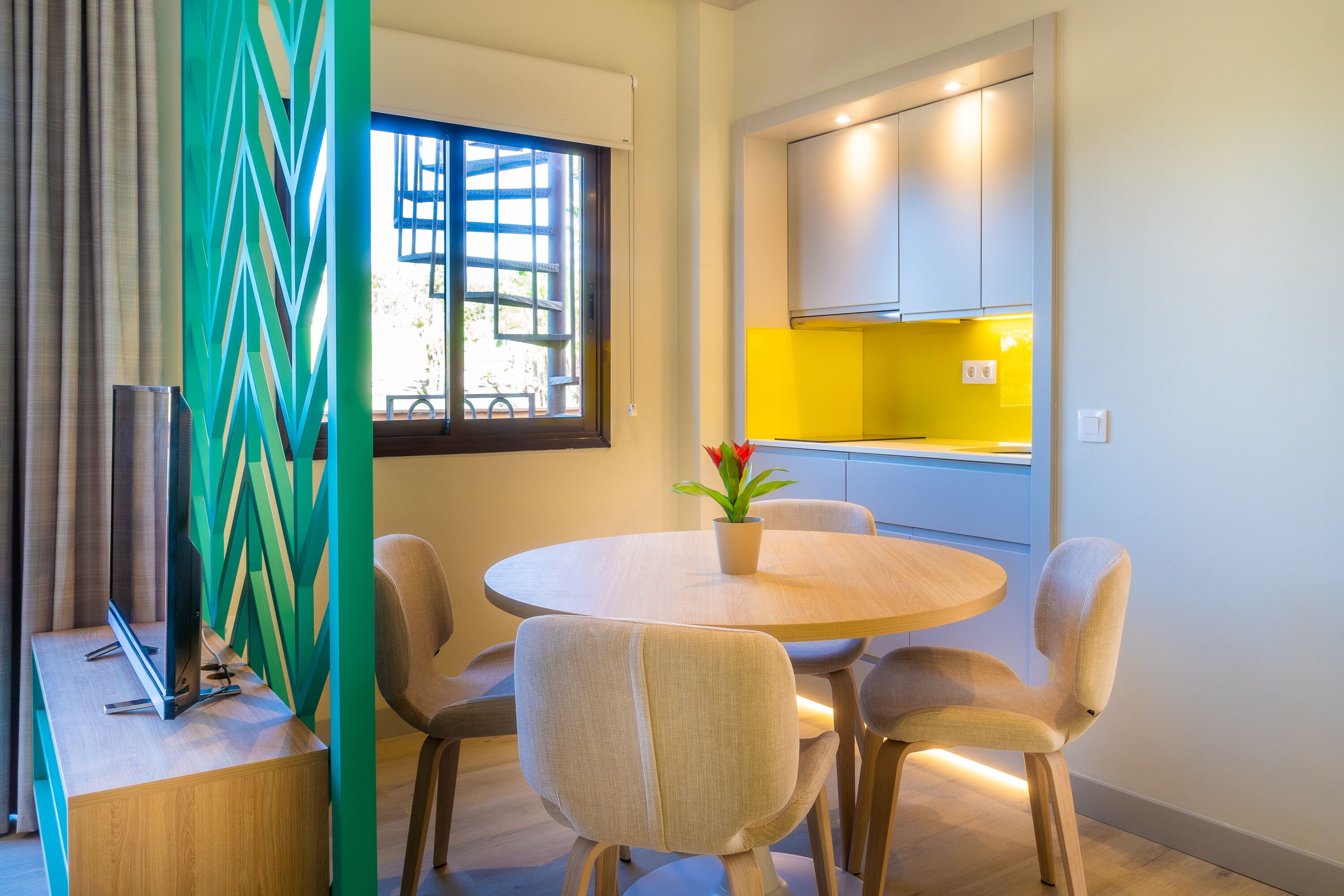 Uma área de refeições moderna com uma mesa redonda e quatro cadeiras, uma planta em vaso e vista para a cozinha.