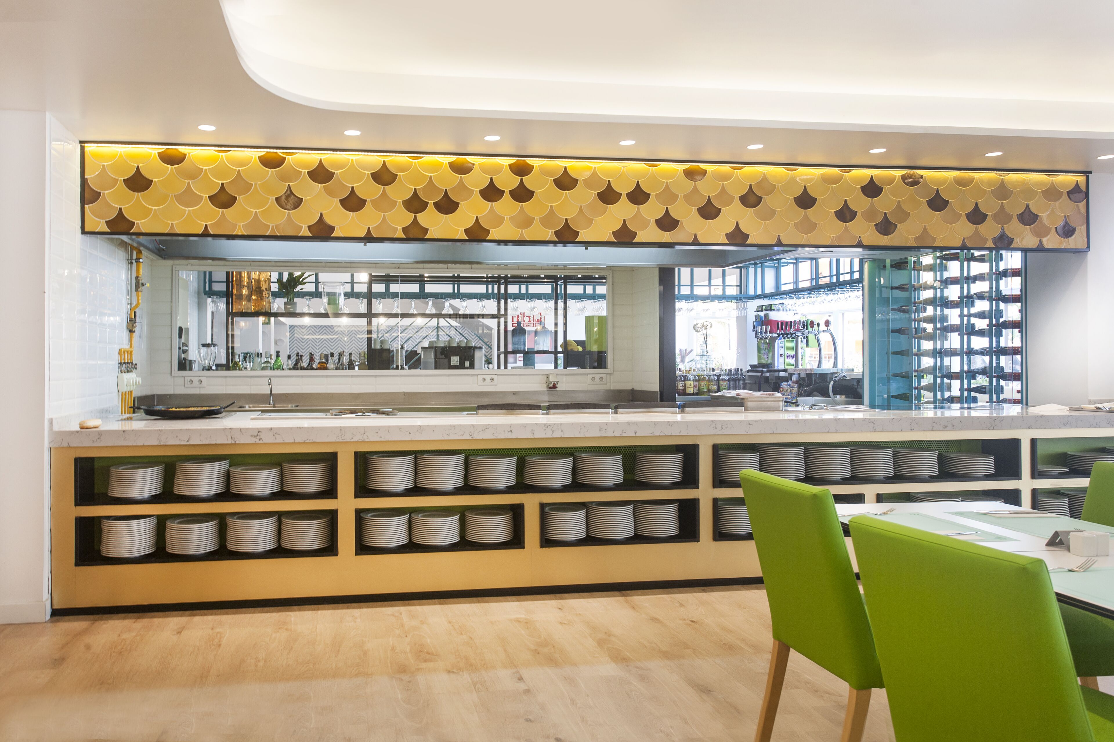 Un restaurante moderno con una barra alargada, sillas verdes y una variedad de platos exhibidos en estanterías.