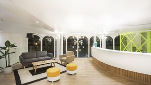 A modern reception area with a curved reception desk, a grey sofa, a wooden coffee table, and two yellow ottomans. - Hotel Villa Mandi Golf Resort (Arona)