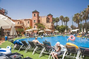 Un área junto a la piscina con sillones de descanso, sombrillas y palmeras.