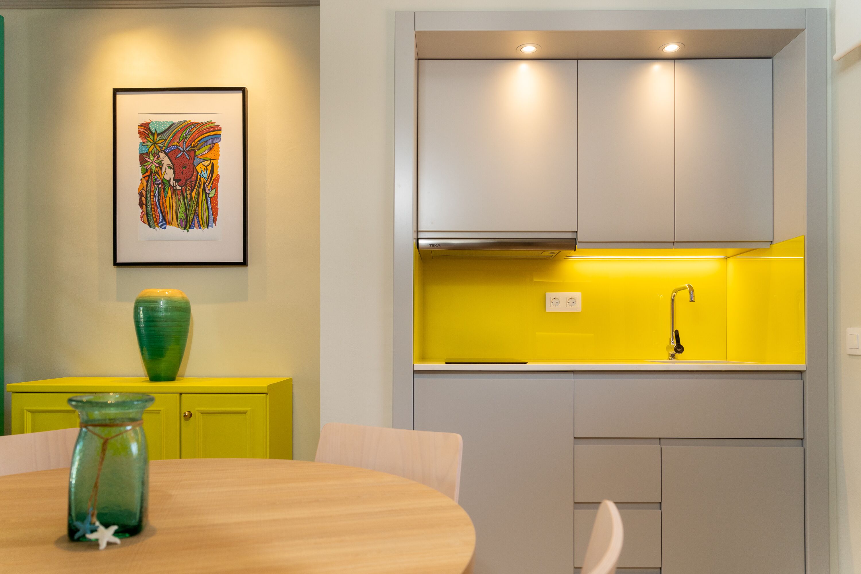 Una cocina moderna con un salpicadero amarillo, una obra de arte enmarcada en la pared y un jarrón verde sobre el mueble amarillo.