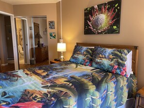 Ein Hotelzimmer mit einem Bett, das mit einem tropischen Bettbezug ausgestattet ist, einem Nachttisch mit Lampe und einem an der Wand befestigten Blumenbild.