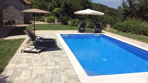 Una piscina rectangular con borde de azulejos, rodeada por un patio de piedra. Hay dos sillas de descanso y un paraguas.