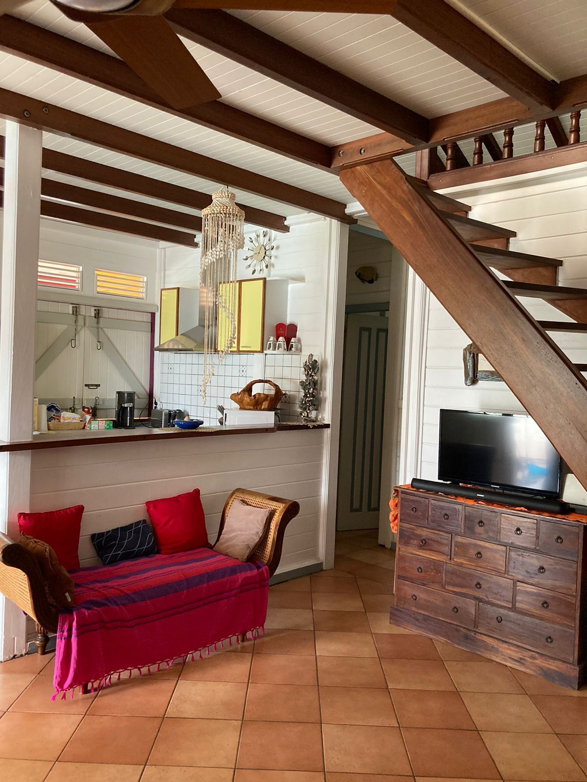 Ein gemütliches Wohnzimmer mit einer Holztreppe, einem Sofa mit bunten Kissen, einem Fernseher auf einem Holzschrank und einer Küchenzeile mit einer Uhr und Dekorationselementen.