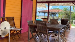 Une terrasse extérieure avec une table et des chaises en bois, un espace couvert avec un toit en métal, et une vue sur un bâtiment avec un toit rouge en arrière-plan.