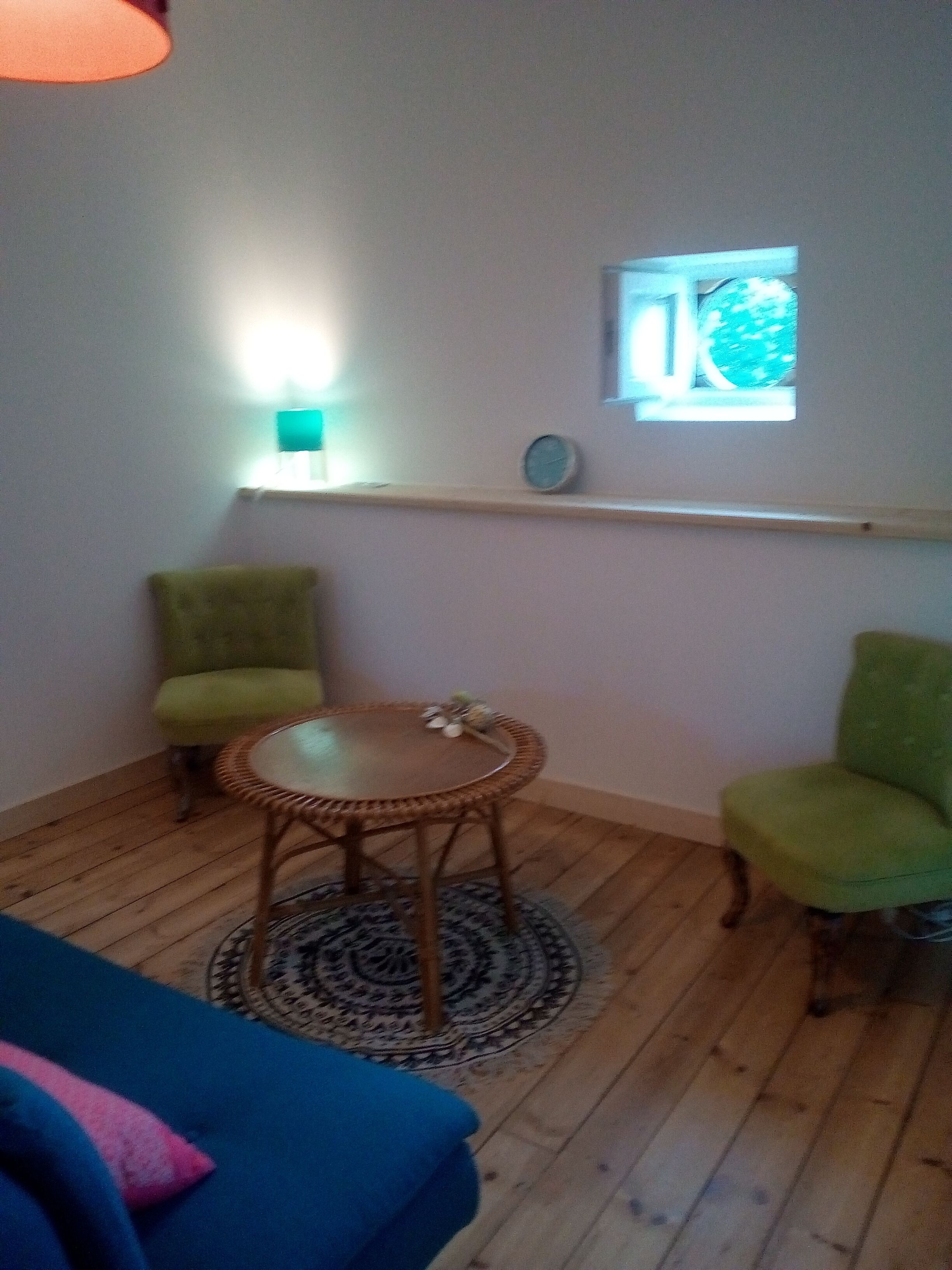 Une pièce avec un plancher en bois, une table ronde en osier, deux fauteuils verts, un canapé bleu, une petite horloge ronde et une fenêtre donnant sur des arbres.