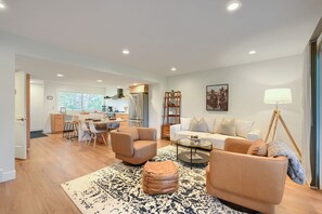 Een moderne woonkamer met een bank, poef en salontafel, een eethoek met stoelen, en een keuken met een koelkast en fornuis.