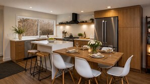 A modern kitchen with a central island, wooden bar stools, white chairs, stainless steel appliances, and a view through the window.