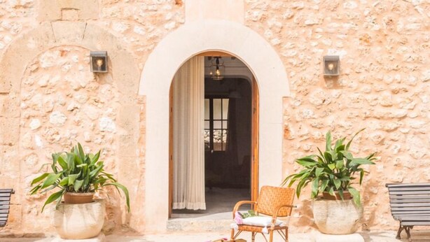 An outdoor seating area with a wicker chair and a bench, flanked by potted plants, in front of a stone building with an arched doorway. - Typical country property with Yacuzzi pool and large outdoor area (Porreres)