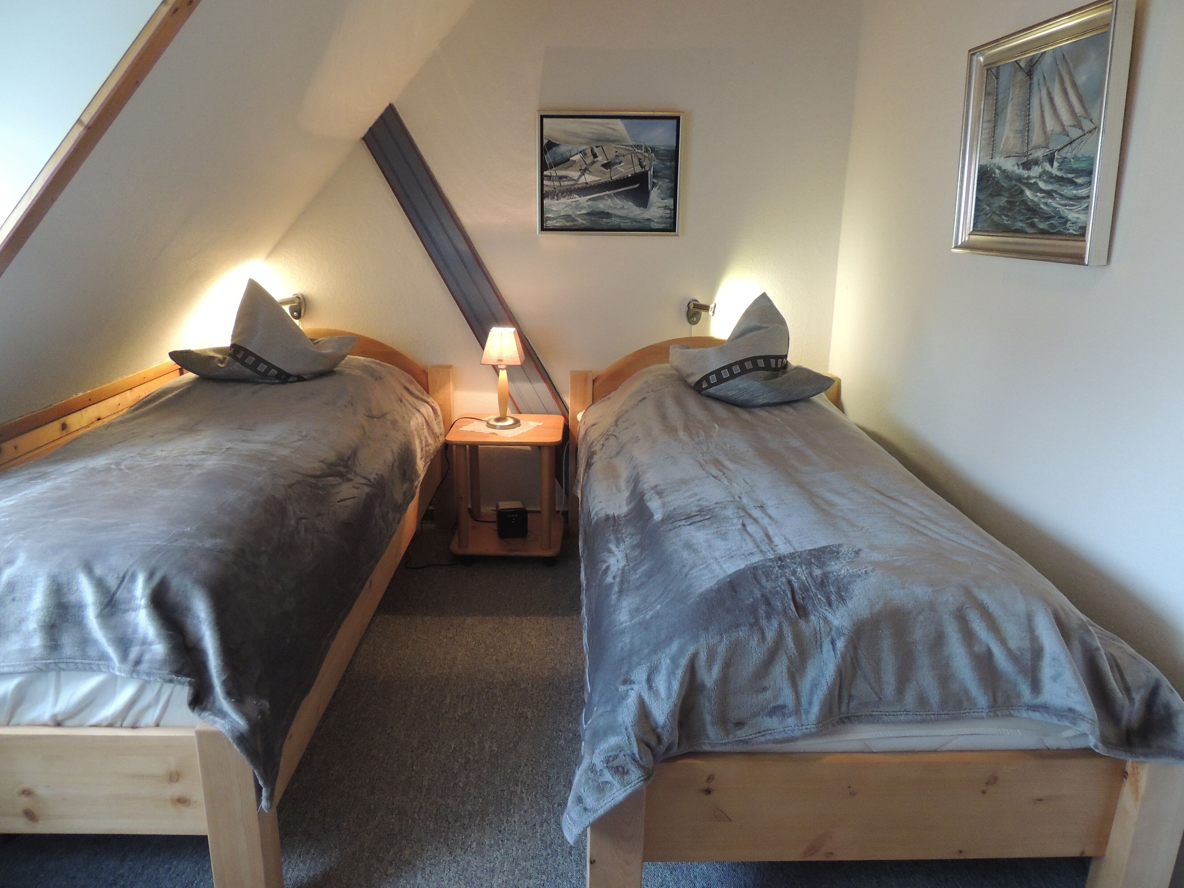 A small, cozy room with two single beds, a nightstand with a lamp, and a painting of a sailboat on the wall.