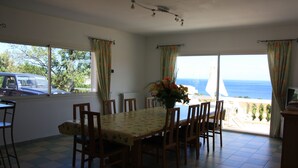 A dining area with a table and chairs, a vase with flowers, and a view of the sea through large windows. - Superb infinity pool villa with stunning sea views in Favone  (Sari-Solenzara)