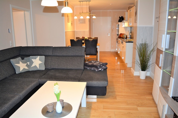Ein modernes Wohnzimmer mit einer grauen Eckcouch, einem weißen Couchtisch mit Blumenvase und einem Essbereich im Hintergrund.
