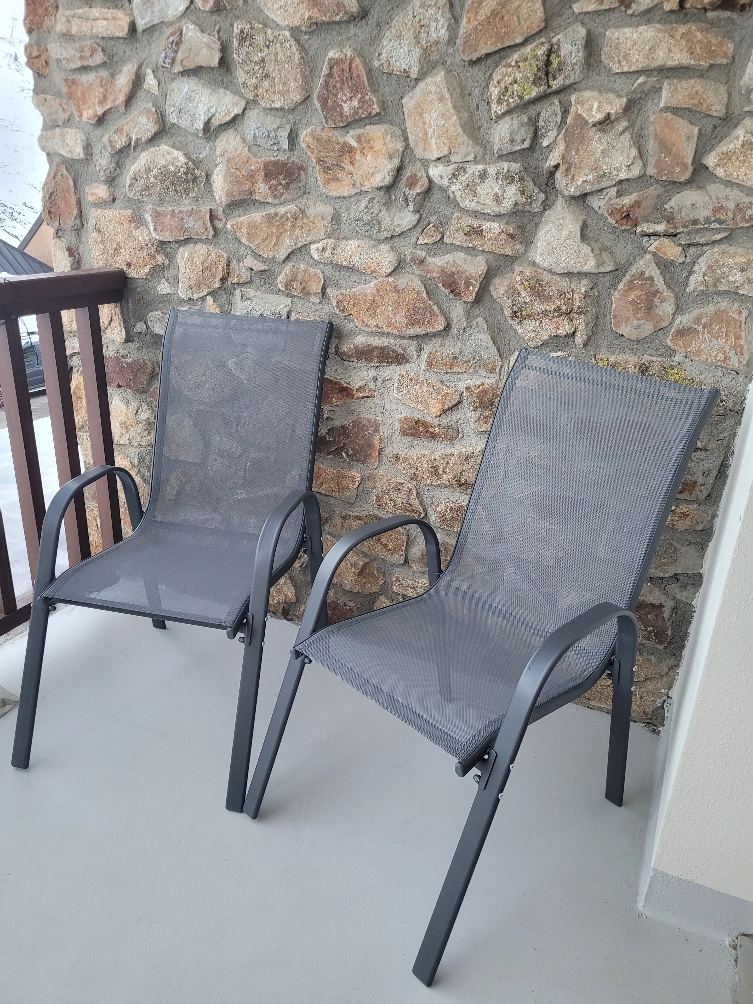 Dos sillas para exteriores con estructura de metal y asientos de malla, ubicadas sobre un patio de concreto frente a una pared de piedra.