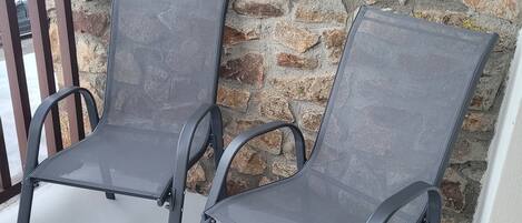 Dos sillas para exteriores con estructura de metal y asientos de malla, ubicadas sobre un patio de concreto frente a una pared de piedra.