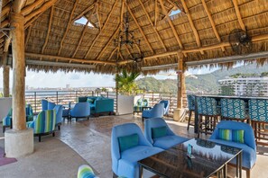 An outdoor seating area with blue chairs and a glass table, overlooking a cityscape and hills. - Residences by Pinnacle, Rooftop Pool & Bar, Zona Romantica, Puerto Vallarta (Puerto Vallarta)