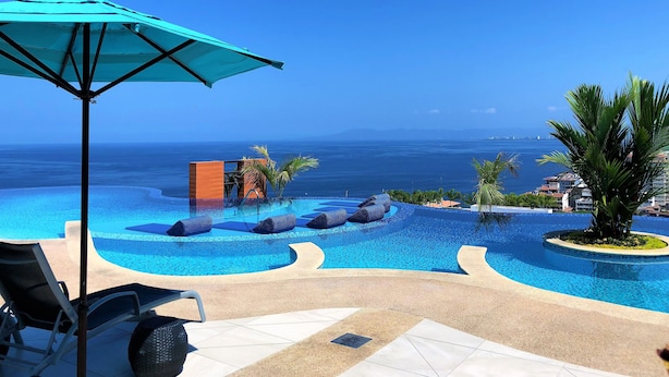 Un área de piscina con sillones y sombrillas, vista al océano y cielo despejado.