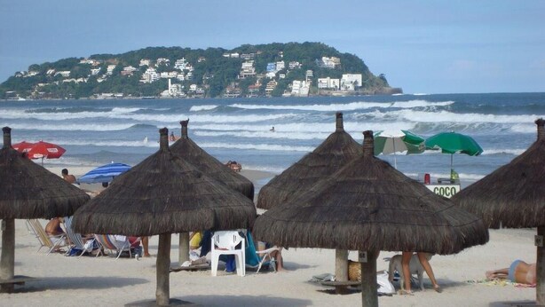 Una playa con sombrillas de paja y vistas a una colina con edificios.