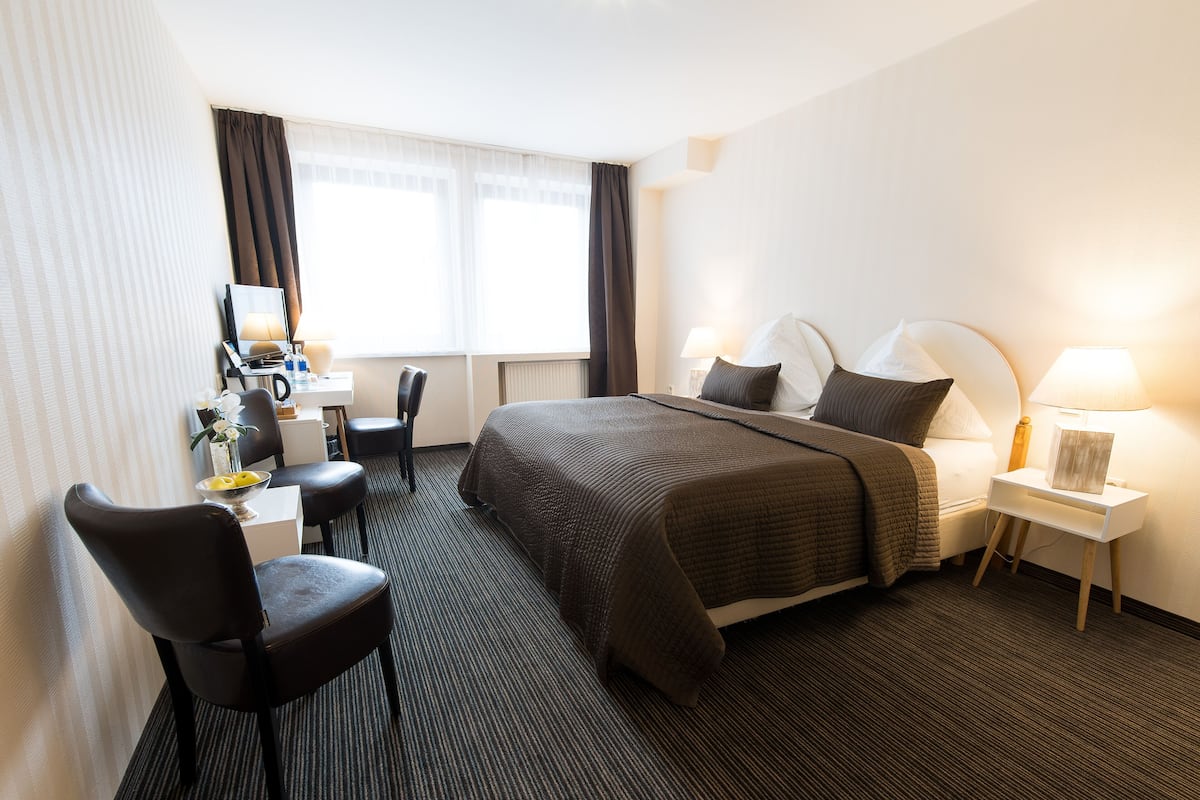 Habitación de hotel con cama, escritorio con silla, lámpara y ventana con cortinas.