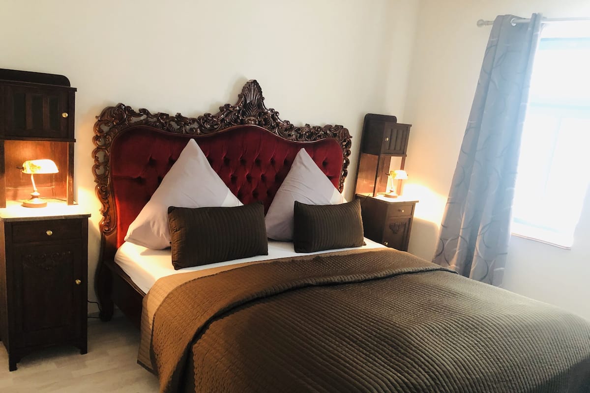 Un dormitorio con cabecera capitonada, cama con varias almohadas, mesitas de noche con lámparas y una ventana con cortinas.